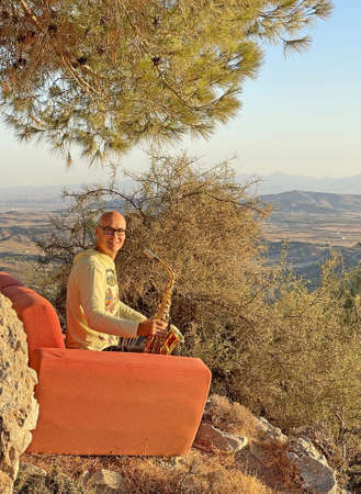 Young stylish bald male saxophonist sits on an orange sofa in mountains of Cyprus in nature, under pine tree, holds an alto saxophone in his hands. Musician with saxophone on nature in mountains.の写真素材
