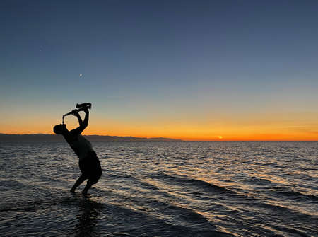 Young male saxophonist stands with his feet in sea water, holds saxophone in his hands, looks at sunset. Beautiful sunset on sea, sky. Musician, playing saxophone, dancing, having fun, Silhouette.の写真素材