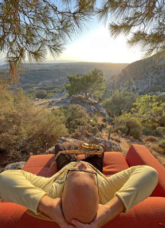 Young stylish bald male saxophonist sits on an orange sofa in mountains of Cyprus in nature, under pine tree, holds an alto saxophone in his hands. Musician with saxophone on nature in mountains.の写真素材