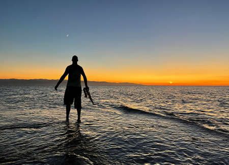 Young male saxophonist stands with his feet in sea water, holds saxophone in his hands, looks at sunset. Beautiful sunset on sea, sky. Musician, playing saxophone, dancing, having fun, Silhouette.の写真素材