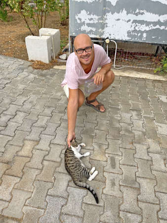 Young bald man gently strokes beautiful gray tabby cat, which lies on street on road, on burgundy mat, outside in summer. Caring, tenderness, affection, kindness, love for animals. Cute beautiful cat.の写真素材