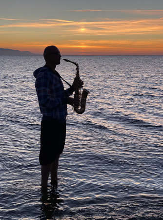 Young male saxophonist stands with his feet in sea water, holds saxophone in his hands, looks at sunset. Beautiful sunset on sea, sky. Musician, playing saxophone, dancing, having fun, Silhouette.の写真素材