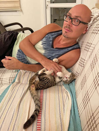 Young bald man gently strokes beautiful gray tabby cat, which lies at home on the couch in the evening. Caring, tenderness, affection, kindness, love for animals. Cute beautiful cat.の写真素材