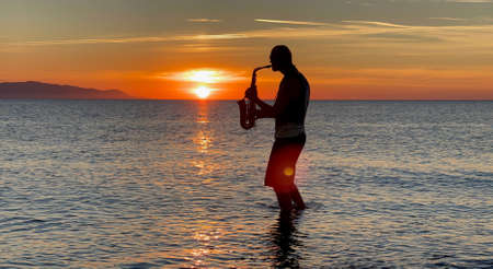 Young male saxophonist stands with his feet in sea water, holds saxophone in his hands, looks at sunset. Beautiful sunset on sea, sky. Musician, playing saxophone, dancing, having fun, Silhouette.の写真素材