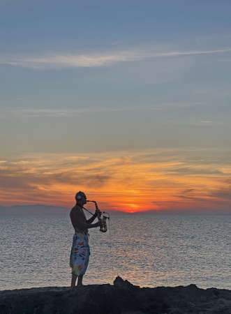 Young male saxophonist stands against background of sea and sunset, holds saxophone in his hands. Beautiful sunset on sea, sky. Musician saxophonist plays against background sea, sunset, silhouette.の写真素材
