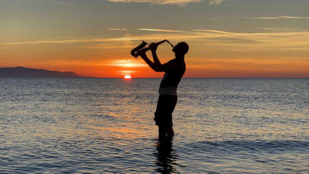 Young male saxophonist stands with his feet in sea water, holds saxophone in his hands, looks at sunset. Beautiful sunset on sea, sky. Musician, playing saxophone, dancing, having fun, Silhouette.の写真素材