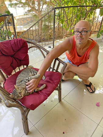 Young bald man gently strokes beautiful gray tabby cat, which lies on soft armchair, on burgundy mat, outside in summer. Caring, tenderness, affection, kindness, love for animals. Cute beautiful cat.の写真素材