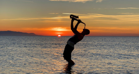 Young male saxophonist stands with his feet in sea water, holds saxophone in his hands, looks at sunset. Beautiful sunset on sea, sky. Musician, playing saxophone, dancing, having fun, Silhouette.の写真素材