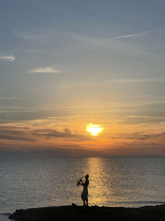 Young male saxophonist stands against background of sea and sunset, holds saxophone in his hands. Beautiful sunset on sea, sky. Musician saxophonist plays against background sea, sunset, silhouette.の写真素材