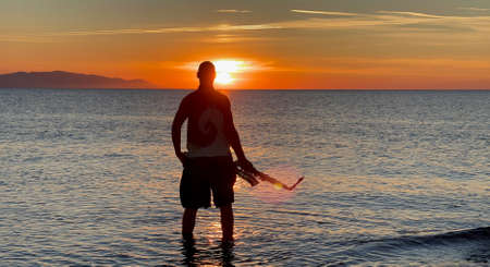Young male saxophonist stands with his feet in sea water, holds saxophone in his hands, looks at sunset. Beautiful sunset on sea, sky. Musician, playing saxophone, dancing, having fun, Silhouette.の写真素材