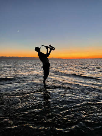 Young male saxophonist stands with his feet in sea water, holds saxophone in his hands, looks at sunset. Beautiful sunset on sea, sky. Musician, playing saxophone, dancing, having fun, Silhouette.の写真素材