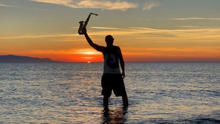 Young male saxophonist stands with his feet in sea water, holds saxophone in his hands, looks at sunset. Beautiful sunset on sea, sky. Musician, playing saxophone, dancing, having fun, Silhouette.の写真素材