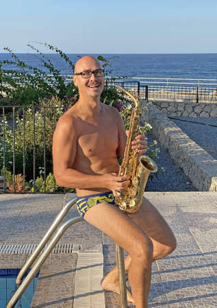 A young male saxophonist in swimming trunks sits on the background of the pool and holds a saxophone musical instrument in his hands and smiles. Handsome saxophonist man on vacation.の写真素材