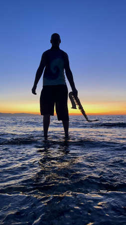 Young male saxophonist stands with his feet in sea water, holds saxophone in his hands, looks at sunset. Beautiful sunset on sea, sky. Musician, playing saxophone, dancing, having fun, Silhouette.の写真素材