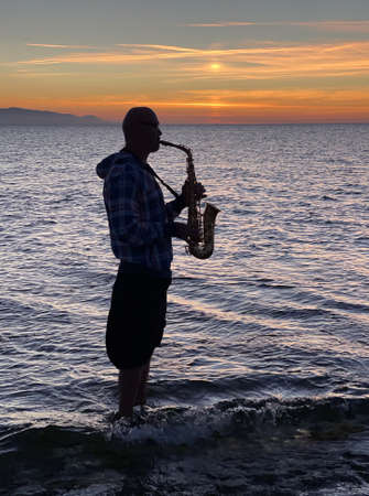 Young male saxophonist stands with his feet in sea water, holds saxophone in his hands, looks at sunset. Beautiful sunset on sea, sky. Musician, playing saxophone, dancing, having fun, Silhouette.の写真素材