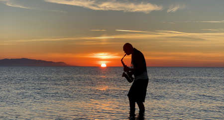 Young male saxophonist stands with his feet in sea water, holds saxophone in his hands, looks at sunset. Beautiful sunset on sea, sky. Musician, playing saxophone, dancing, having fun, Silhouette.の写真素材