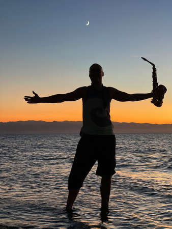 Young male saxophonist stands with his feet in sea water, holds saxophone in his hands, looks at sunset. Beautiful sunset on sea, sky. Musician, playing saxophone, dancing, having fun, Silhouette.の写真素材