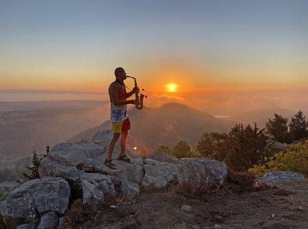 Young stylish bald male saxophonist stands on top of mountain against the backdrop of the sunset and the sun and plays the golden saxophone in Cyprus. Romantic Musician saxophonist. Above clouds.の写真素材