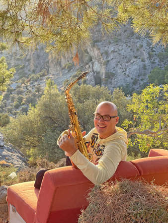 Young stylish bald male saxophonist sits on an orange sofa in mountains of Cyprus in nature, under pine tree, holds an alto saxophone in his hands. Musician with saxophone on nature in mountains.の写真素材