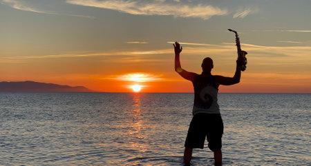 Young male saxophonist stands with his feet in sea water, holds saxophone in his hands, looks at sunset. Beautiful sunset on sea, sky. Musician, playing saxophone, dancing, having fun, Silhouette.の写真素材