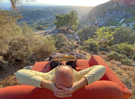 Young stylish bald male saxophonist sits on an orange sofa in mountains of Cyprus in nature, under pine tree, holds an alto saxophone in his hands. Musician with saxophone on nature in mountains.の写真素材