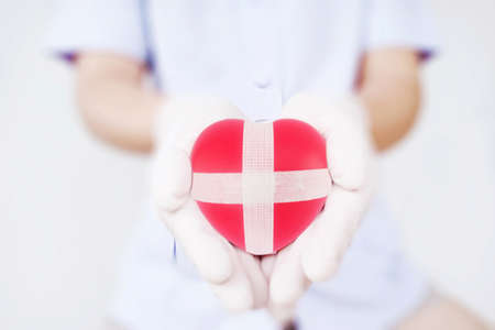 The doctor wearing white shirt with gloves holding a red heart adhesived with white plaster with over bright and blur area surronding the red heart.の写真素材