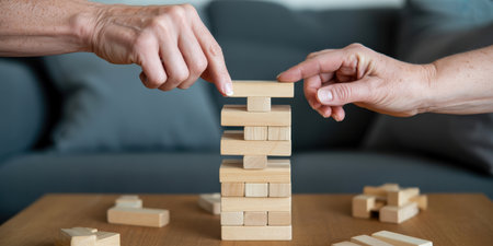 Close-up view of two hands, one appearing older and the other younger, carefully placing wooden blocks onto a Jenga tower. The blocks are light brown wood, and the tower is partially built on a wooden table. In the background, a dark grey sofa is visible, slightly out of focus. Scattered wooden blocks are also on the table.の素材