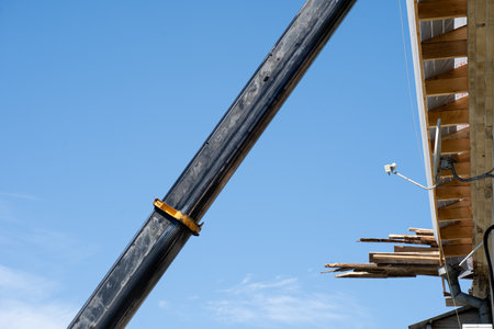 A large crane is positioned at a construction site, lifting wooden beams toward the roof. The bright...