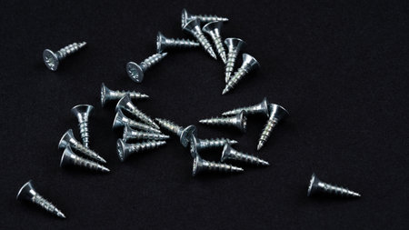 A collection of small metal screws is scattered across a black surface, highlighting their shiny texture and sharp tips. These screws vary slightly in size, useful for various crafts and repairs.の写真素材