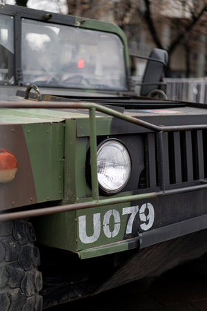 A military vehicle is stationed on a city street, featuring a distinctive green and brown camouflage design. The weather is cloudy, creating a dramatic atmosphere around the rugged transport.の写真素材