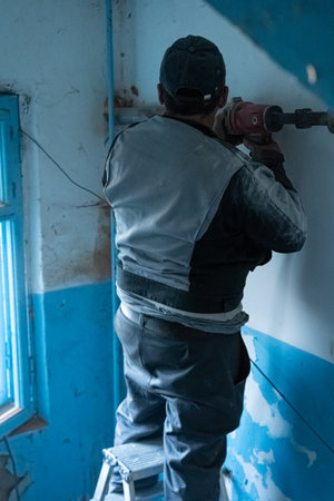 A construction worker is using a power drill to make holes in a wall of an old building. The worker is wearing a safety vest and standing on a stool while surrounded by blue and white paint.の写真素材