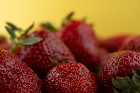 Juicy strawberries glisten under soft light, showing their vibrant red color and tiny seeds, with lush green leaves contrasting against a bright yellow backdrop.の写真素材
