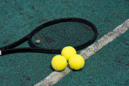 A black tennis racket rests on a green court next to three yellow tennis balls. The court surface shows signs of wear, suggesting frequent use, ideal for practice or casual play.の写真素材
