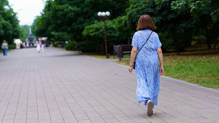 A woman strolls along a paved path in a serene park, wearing a blue dress. The atmosphere is peaceful, with trees lining the walkway and soft sunlight filtering through the leaves.の写真素材