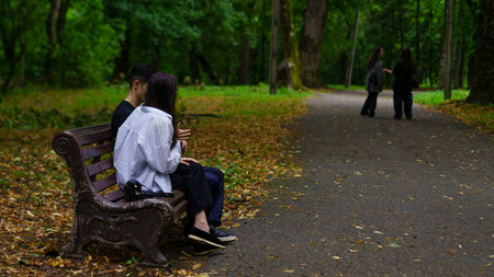 In a serene park, a couple sits closely on a bench, embracing the moment as fallen leaves cover the ground. Friends stroll in the background, creating a lively atmosphere.の写真素材