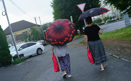 Two women are strolling down a city street, carrying red bags and using colorful umbrellas as it rains. The atmosphere is calm with greenery around and cars parked nearby.の写真素材