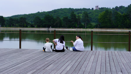 Three individuals sit on a wooden dock overlooking a calm lake, enjoying quality time together. The atmosphere is relaxed, surrounded by greenery and soft overcast skies.の写真素材