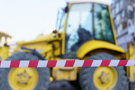 A construction site features a yellow backhoe loader partially obscured by safety tape. The machinery is set against an urban backdrop, suggesting ongoing development.の写真素材