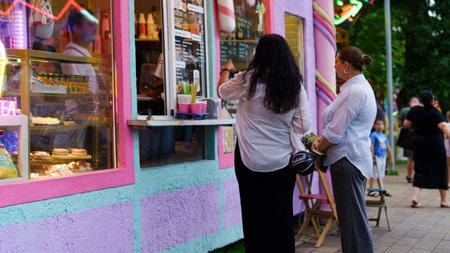 Two women stand in front of a vibrant food stand, placing their orders as colorful lights illuminate the evening atmosphere in a busy park.の写真素材