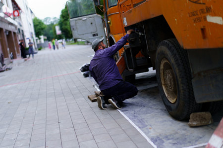 A mechanic is crouching near a large orange truck, working diligently on repairs in a lively city plaza filled with pedestrians during the day.の写真素材