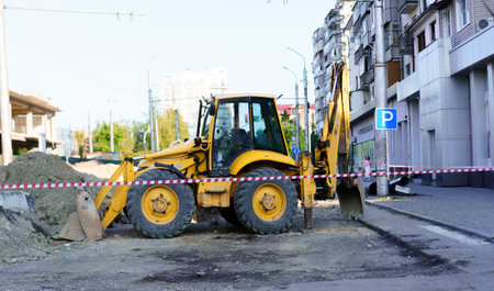 Heavy machinery is parked on a city street where construction and repairs are taking place. Safety barriers are set up around the ongoing work area in broad daylight.の写真素材