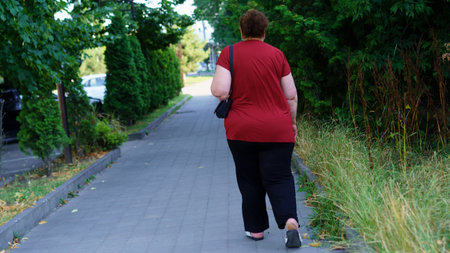 A person in a red shirt and black pants walks peacefully along a sidewalk bordered by trees and tall grass on a sunny day in a quiet suburban neighborhood.の写真素材
