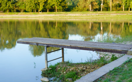 A rustic wooden dock reaches out into a tranquil lake reflecting the surrounding trees under bright daylight. The peaceful atmosphere invites relaxation and nature appreciation.の写真素材