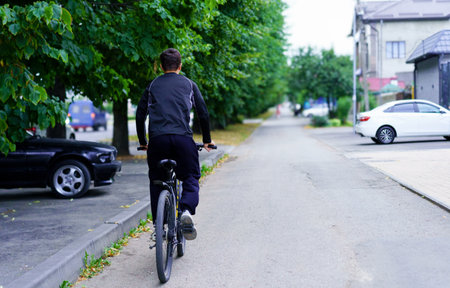 A person in casual attire cycles along a peaceful, green-lined road surrounded by homes and parked cars. The atmosphere is calm and serene, reflecting a typical day.の写真素材
