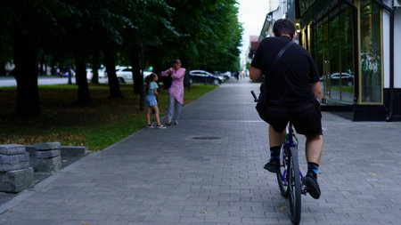 A man rides his bicycle down a quiet street lined with trees during the evening. Two people stand by a store, engaged in conversation, while cars are parked along the road.の写真素材