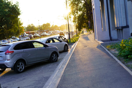 Sunlight casts a warm glow on a quiet street as parked cars line the side of the road. A pedestrian path runs adjacent, offering a serene evening atmosphere.の写真素材