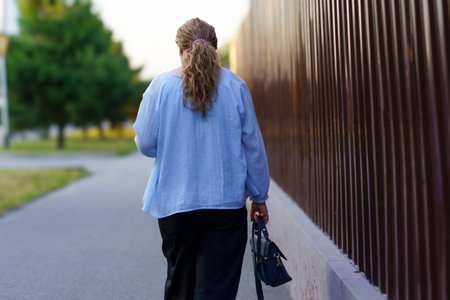 A woman strolls along a peaceful pathway, holding a handbag, as the sun sets behind her. The calm atmosphere and warm light create a serene environment in this urban park.の写真素材