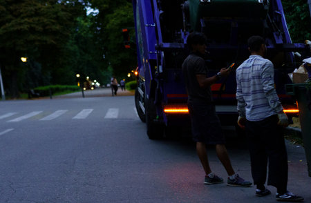 Two workers engage in waste collection near a parked garbage truck while pedestrians stroll in the background during the early evening in a serene park.の写真素材