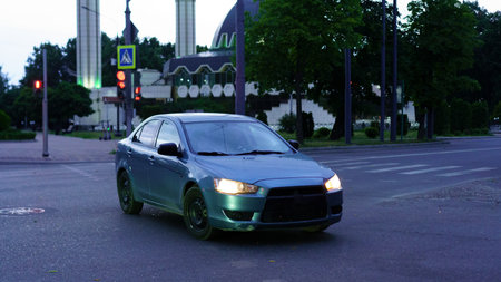 A blue sedan is seen driving on an intersection as streetlights glow softly in the background. The scene captures the essence of an evening in an urban area with notable buildings.の写真素材