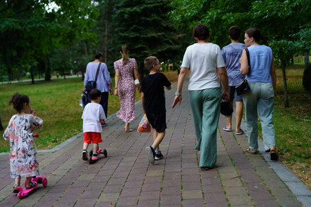 A joyful group walks along a path in a lush green park during the evening. Children ride scooters ahead, while adults share conversation and laughter in a relaxed atmosphere.の写真素材
